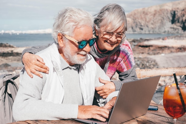 Glückliches älteres Paar, das zusammen auf dem Laptop surft und den sonnigen Tag im Freien am Strand genießt