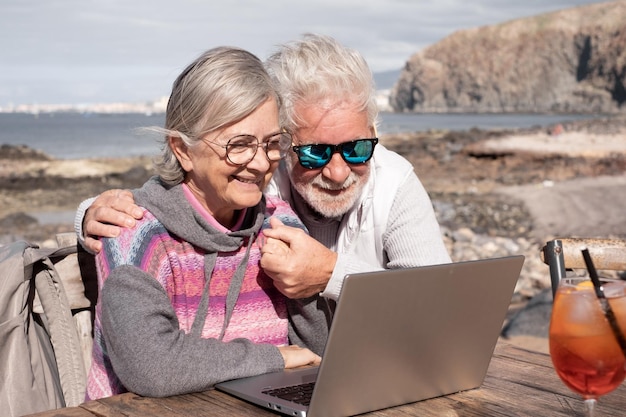 Glückliches älteres Paar, das zusammen auf dem Laptop surft und den sonnigen Tag im Freien am Strand genießt