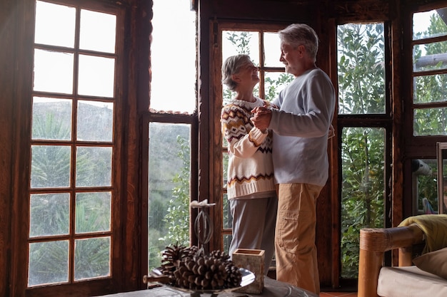 Glückliches älteres Paar in Liebe tanzt zu Hause gegen eine große Fensteransicht mit Naturwald draußen. Romantik Freizeitbeschäftigung reifer Mann und Frau älterer Lebensstil im Ruhestand