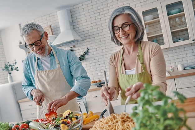 Glückliches älteres Paar in Schürzen, das gesundes Abendessen zubereitet und lächelt, während es Zeit zu Hause verbringt