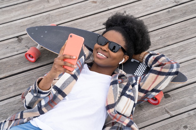 Glückliches afrikanisches Mädchen mit Longboard-SMS im Smartphone, das auf dem Boden im städtischen Raum auf Skateboard liegt