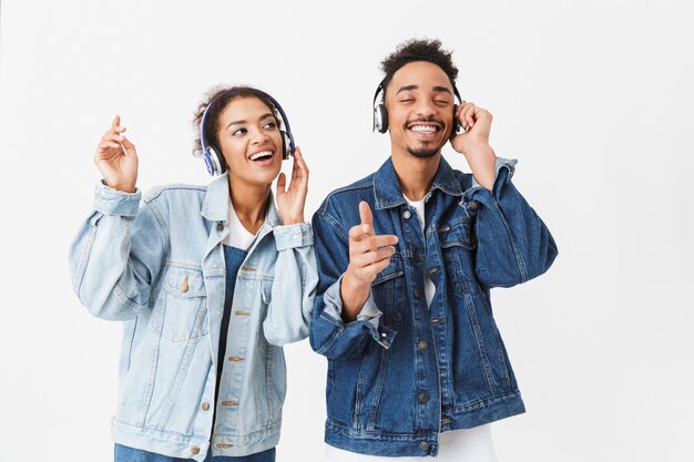 Glückliches afrikanisches Paar in Jeanshemden, die Musik durch Kopfhörer hören und über graue Wand tanzen