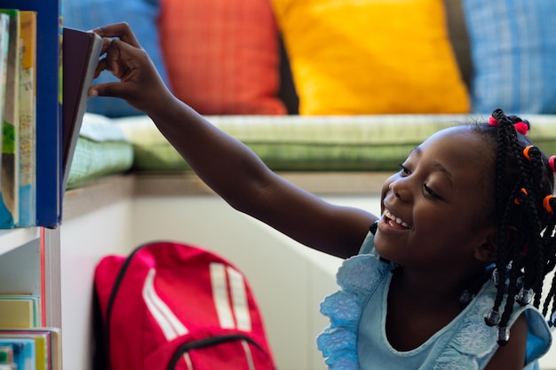 Glückliches afroamerikanisches Schulmädchen, das ein Buch in einem bunten Raum in der Grundschule auswählt