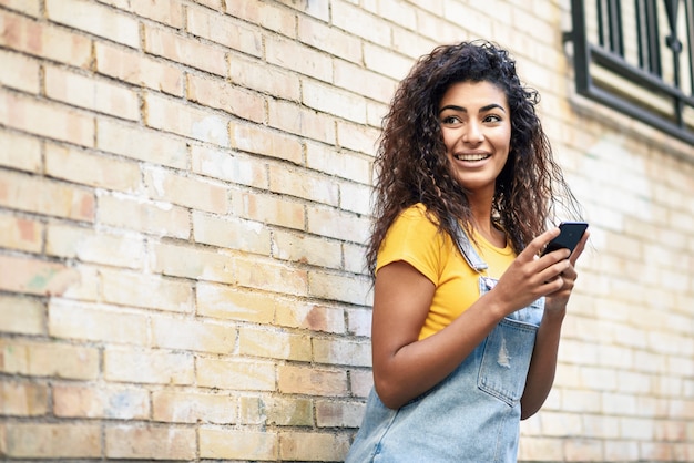 Glückliches arabisches Mädchen, das intelligentes Telefon auf Backsteinmauer verwendet.