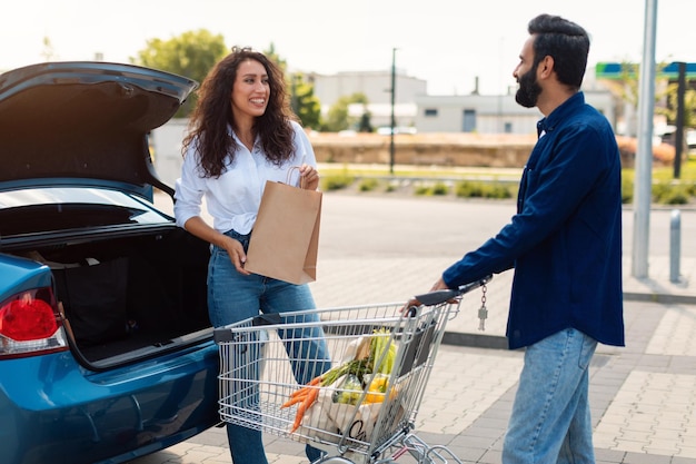 Glückliches arabisches Paar, das Einkaufstüten in den Kofferraum eines Autos packt, während eine Dame auf einem Parkplatz schaut und redet