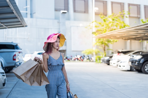 Glückliches asiatisches beiläufiges Kleid der Frau in Mode mit Einkaufstaschen und Gehen in Parkplatz.