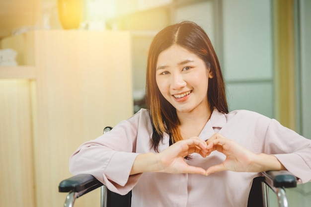 Glückliches asiatisches jugendlich Mädchenlächeln Herzformhand, die auf Rollstuhl im Krankenhaus sitzt