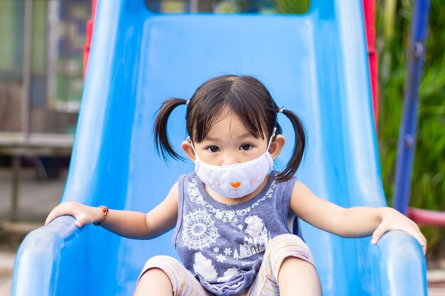 Glückliches asiatisches Kindermädchen lächelt und trägt Stoffmaske, Sie spielt mit Schieberegler Spielzeug am Spielplatz.