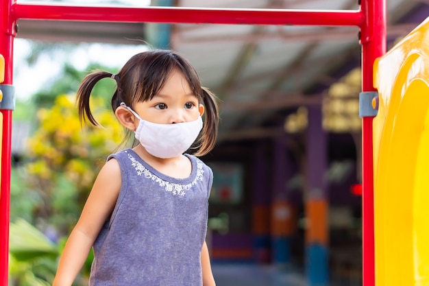 Glückliches asiatisches Kindermädchen lächelt und trägt Stoffmaske, Sie spielt mit Schieberegler Spielzeug am Spielplatz.