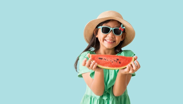 Glückliches asiatisches kleines Mädchen, das einen Hut mit Sonnenbrille trägt und Wassermelonenscheiben in der Hand hält. Urlaubs-Sommermode Grünes Kleid isoliert auf pastellblauem Hintergrund