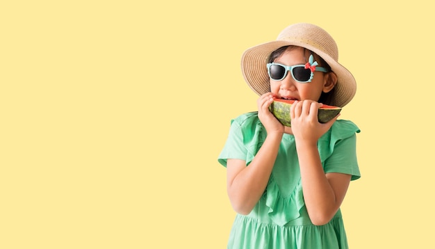 Glückliches asiatisches kleines Mädchen, das einen Hut mit Sonnenbrille trägt und Wassermelonenscheiben in der Hand hält. Urlaubs-Sommermode Grünes Kleid isoliert auf pastellgelbem Hintergrund