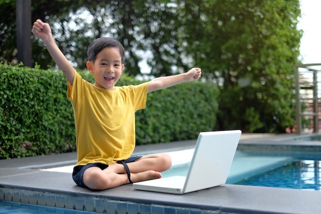 Glückliches asiatisches Kleinkind, das Computerlaptop mit der Hand oben an der Seite des Swimmingpools verwendet