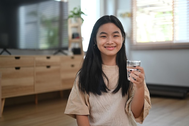 Glückliches asiatisches Mädchen, das ein Glas Wasser hält und vor der Kamera lächelt. Gesunder Lebensstil-Konzept.