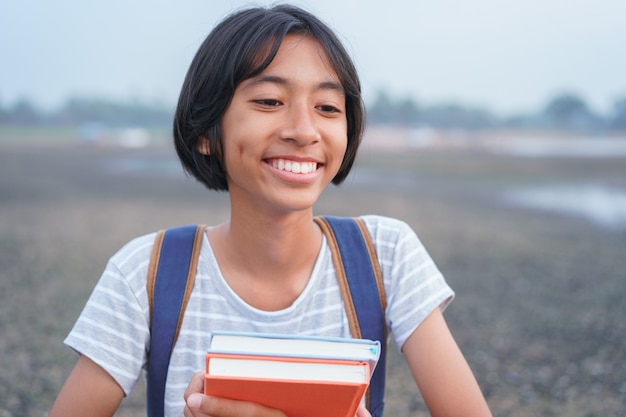 Glückliches asiatisches Mädchenlächeln auf Gesicht und lachen, während am Morgen inmitten der Natur Asien-Kind Buch und Rucksack halten