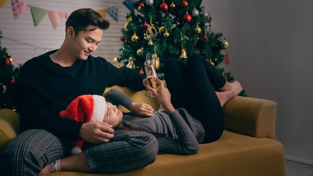 Glückliches asiatisches Paar, das zusammen Champagner anstößt, sitzt abends auf dem Sofa mit einem Weihnachtsbaum und Lichtern im Hintergrund Freund und Freundin in der Heiligabendnacht