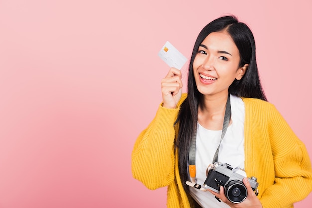 Glückliches asiatisches Porträt, schöne junge Frau, aufgeregter lächelnder Fotograf, der eine Kreditkarte und eine Retro-Vintage-Fotokamera hält, die bereit ist, auf rosa Hintergrund, Tourismus und Urlaub zu reisen