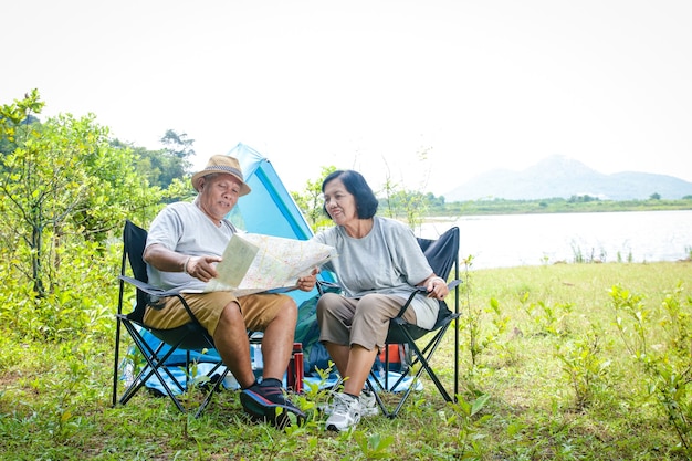 Glückliches asiatisches Seniorenpaar Camping am Wasser, auf einem Stuhl sitzend, touristische Karte anzeigen, mit Kopierraum.