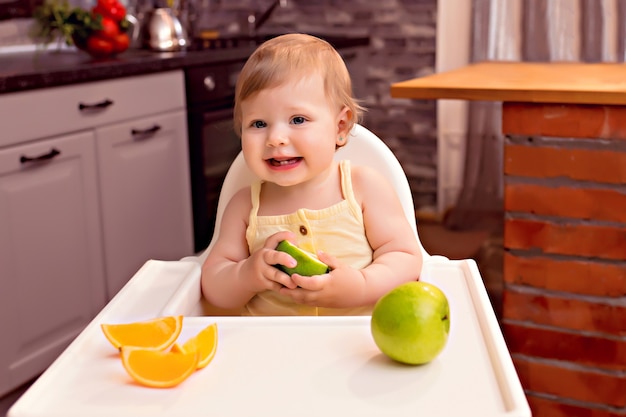 Glückliches Baby 10-12 Monate isst Obst: Orange, Apfel. Porträt eines glücklichen Mädchens in einem Hochstuhl in der Küche