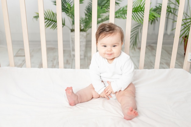 Glückliches Baby, das in einem Kinderbett in einem weißen Bodysuit sitzt