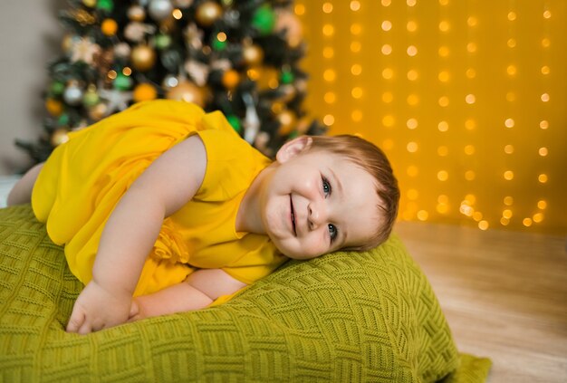 Glückliches Baby in einem gelben Kleid, das auf einem grünen Kissen mit Weihnachtsbaum liegt