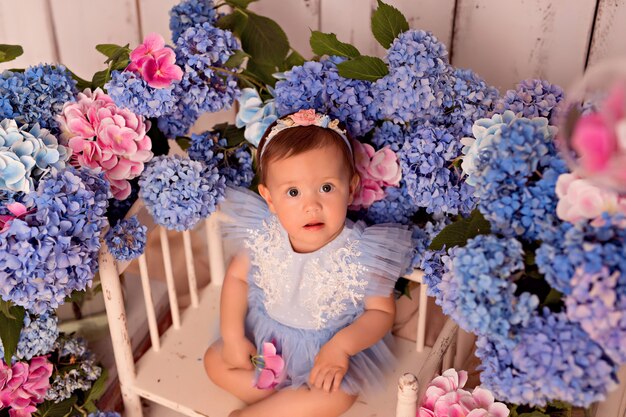 Glückliches Baby in einem Kleid im Studio auf einem weißen Hintergrund mit rosa und blauen Hortensienblumen