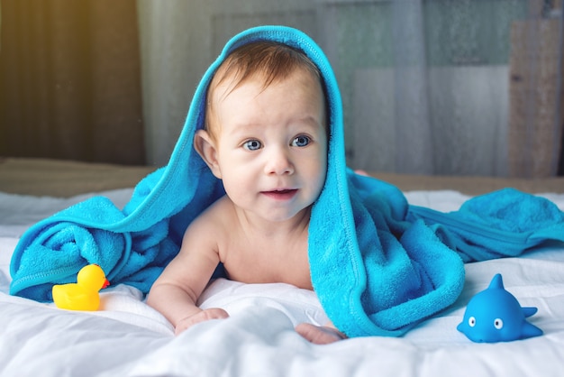 Glückliches Baby mit blauen Augen, die auf einer Decke mit einer Ente liegen, die nach dem Baden in ein Handtuch gewickelt wird