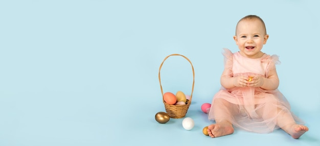 Glückliches Baby, zehn Monate alt, trägt Hasenohren Stirnband, das auf hellblauem Studiohintergrund sitzt und mit farbigen Ostereiern spielt Kleines Kind Ostergrußkarte Baby-Osterhase