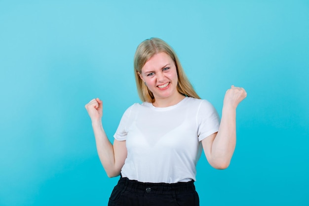 Glückliches blondes Mädchen erhebt ihre Fäuste auf blauem Hintergrund