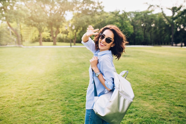 Glückliches brünettes Mädchen mit kurzen Haaren in Sonnenbrille geht mit Tasche im Park spazieren. Sie trägt weißes T-Shirt, Hemd und Jeans. Sie lächelt in die Kamera.