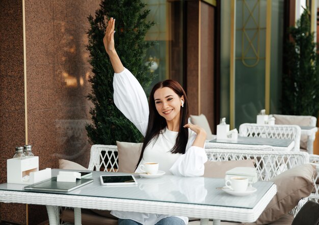 Glückliches brünettes Mädchen sitzt in einem Café und winkt einem Freund auf der Straße mit der Hand.