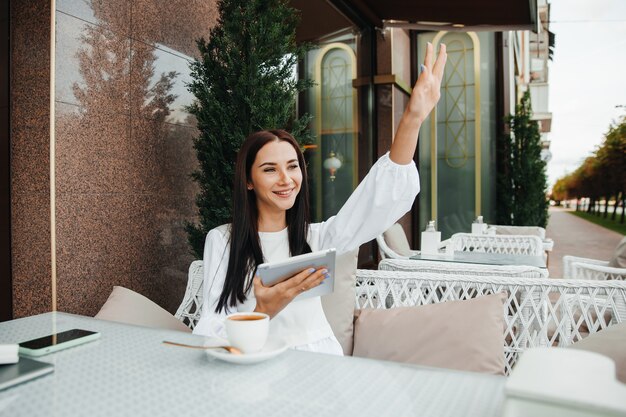 Glückliches brünettes Mädchen sitzt in einem Café und winkt einem Freund auf der Straße mit der Hand.