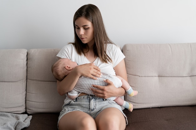 Glückliches Elternkonzept. Junge schöne Frau sitzt auf einem Sofa. Halten Sie ein kleines Kind in den Armen