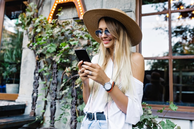 Glückliches entzückendes Frau-Scrolling-Smartphone mit fröhlichem Lächeln im Freien gekleidetes Hemd und Shorts, Hut und stilvolle Sonnenbrille, glückliches Lächeln, Sommer