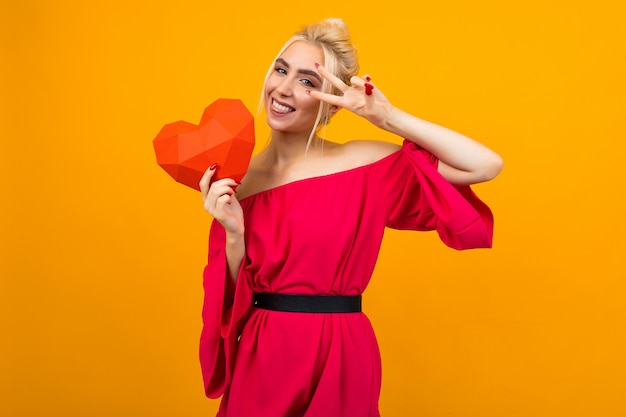 Glückliches freudiges lächelndes blondes Mädchen im roten Kleid hält rotes Papierherz