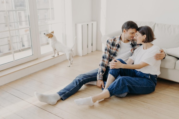 Glückliches frisch verheiratetes Paar, das in eine neue Wohnung zieht, freut sich, gemeinsam Freizeit zu haben, umarmt und berührt Nasen, die süße Kuss-Pose auf dem Boden in einem leeren Raum in der Nähe des Sofas haben, ihren Hund im Hintergrund