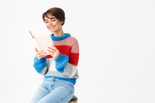 Glückliches fröhliches Mädchen, das Pullover trägt, sitzt auf einem Stuhl lokalisiert auf Weiß, liest ein Buch