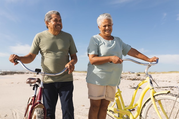 Glückliches, gemischtrassiges älteres Paar, das an sonnigen Tagen mit Fahrrädern am Strand gegen den Himmel steht