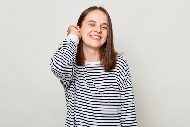 Glückliches Gesicht, positive Emotionen. Lächelnde, attraktive, fröhliche Frau mit braunen Haaren, die ein gestreiftes Hemd trägt und isoliert vor grauem Hintergrund posiert