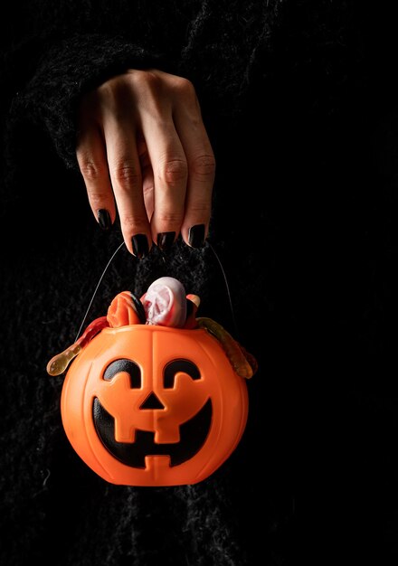 Foto glückliches halloween-konzept. gruselige frau halloween-hand mit schwarzen nägeln, die kürbis voller süßigkeiten auf dunklem hintergrund mit kopienraum hält