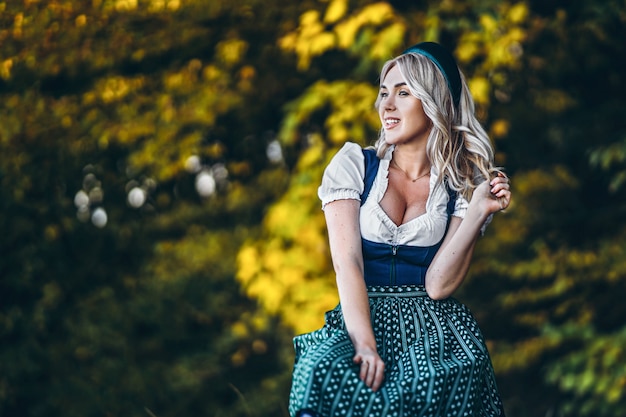 Glückliches hübsches blondes Mädchen in dirndl, traditionellem Bierfestkleid, draußen sitzend mit blutigen bunten Bäumen dahinter