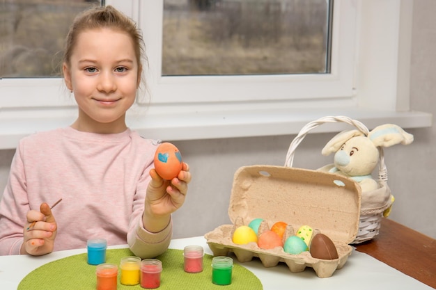 Glückliches hübsches Mädchen, das an einem Tisch mit Farben sitzt, und ein Osterhase zeigt ein bemaltes Osterei