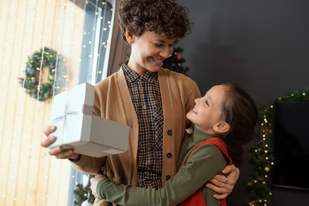 Glückliches hübsches Mädchen, das Mutter mit Geschenkbox umarmt, während sie ihr zu Weihnachten gratuliert