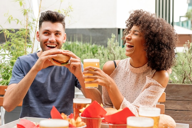 Glückliches hungriges multikulturelles Paar, das Hamburger und kaltes Bier in einem Restaurant oder Pub im Freien genießt und glücklich über einen guten Witz lacht