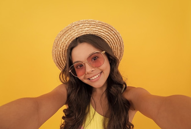 Glückliches jugendlich Mädchen im Sommerstrohhut und -brille, die selfie auf gelber Hintergrundkindheit machen