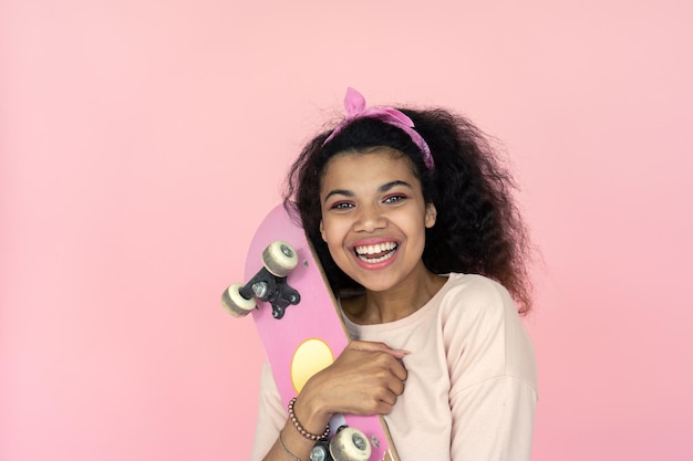 Glückliches junges afrikanisches Mädchen lacht und hält ein Skateboard auf rosa Hintergrund.