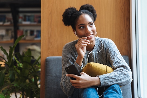 Glückliches junges afrikanisches Studentenmädchen, das an der Bibliothek studiert und Musik mit Kopfhörern hört