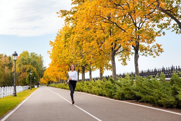 Glückliches junges erwachsenes Mädchen, das im Herbstpark joggt, Kamera und zahniges Lächeln betrachtet. Draussen