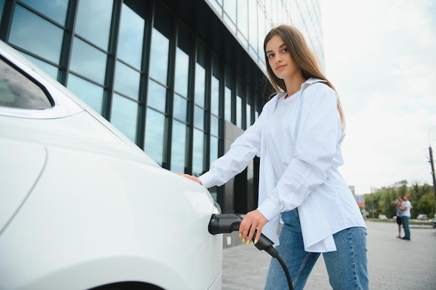 Glückliches junges erwachsenes Mädchen, das Stromkabel in der Hand hält und in der Nähe der öffentlichen Ladestation für Elektroautos steht