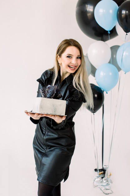 Glückliches junges Geburtstagskind im schwarzen Lederkleid, lächelnd, weißen quadratischen Kuchen haltend, auf dem weißen Hintergrund stehend. Große Luftballons, blaue, weiße und schwarze Farbe, im Hintergrund