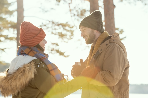 Glückliches junges liebevolles Paar in warmer Winterkleidung mit heißem Tee, während sie voreinander stehen, reden und das Winterwochenende genießen